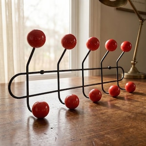 May include: A black metal coat rack with seven red ball-shaped hooks. The rack is mounted on a wooden surface, with a window and lamp in the background. The hooks are evenly spaced and have a glossy finish.