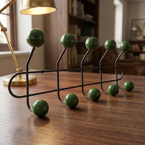 Könnte beinhalten: Ein schwarzer Metall-Wandgarderobenständer mit sechs dunkelgrünen kugelförmigen Knöpfen. Der Ständer hat eine horizontale Stange mit gebogenen Enden und vertikalen Halterungen für die Knöpfe. Die Knöpfe sind gleichmäßig verteilt und glänzend.