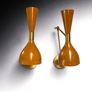 May include: A pair of mid-century wall sconces. Each light has a mustard-yellow, bell-shaped shade with brass detailing. The sconces have a conical top and a small, round base. One sconce has an adjustable arm, and both are mounted on the wall.