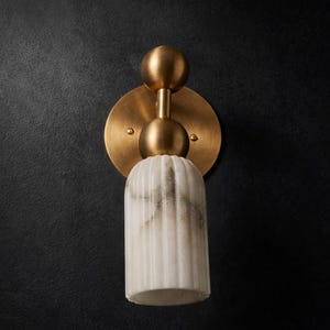 Può includere: Un'applique da parete con finitura in ottone e paralume in marmo bianco. L'apparecchio ha una piastra posteriore circolare, due accenti sferici e un paralume cilindrico con scanalature verticali. La luce è montata su una parete nera.