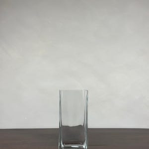 May include: A clear, rectangular glass vase. The vase has a square base and a tall, slender body. It is on a dark brown wooden surface against a light grey wall.