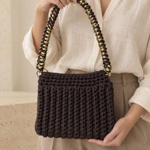 Può includere: Una borsa a mano rettangolare all'uncinetto color marrone scuro con un motivo a maglia spesso e strutturato. La borsa presenta un manico intrecciato che combina corda marrone scuro e accenti in oro metallizzato, fissato con anelli dorati.
