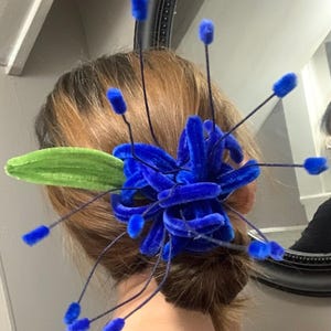May include: A vibrant blue velvet hair accessory, resembling a stylized flower, is secured in the hair. The design includes a cluster of blue petals and several thin stems with blue accents. A green leaf adds a contrasting element.