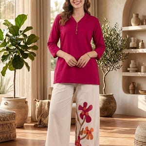 May include: A woman wearing a magenta tunic with three-quarter sleeves and cream-colored palazzo pants. The pants feature a floral design in shades of red, orange, and brown. The outfit is styled with white sandals.