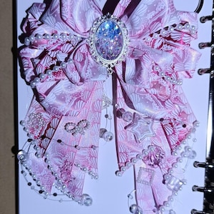 Può includere: Un grande fiocco di nastro rosa con un motivo floreale e dettagli di perle. Il fiocco presenta un elemento centrale ovale argentato pieno di glitter. Ornamenti chiari a forma di stella e altri pendenti decorativi pendono dal fiocco.