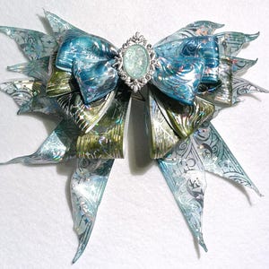 Può includere: Un grande fiocco ornato con strati di nastri blu, verdi e argentati. Il fiocco presenta un abbellimento centrale di forma ovale con una pietra azzurra e una cornice decorativa argentata. I nastri hanno una qualità brillante e iridescente.