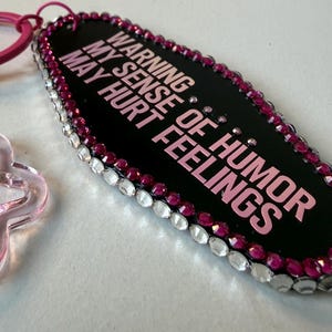 May include: Black keychain with the text "WARNING MY SENSE OF HUMOR MAY HURT FEELINGS" in pink. The keychain is outlined with pink and clear rhinestones and has a pink ring and clasp.