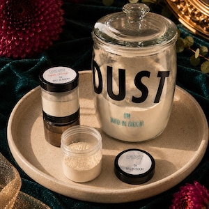 May include: A large clear glass jar with a lid labelled DUST BY WILD IN BLOOM, accompanied by two smaller clear plastic jars with black lids. One small jar is labelled DUST SWEET ORANGE BY WILD IN BLOOM, and the other contains a light beige powder.