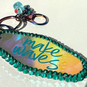 Puede incluir: Llavero iridiscente con las palabras "make waves" en escritura turquesa. El llavero tiene un borde de gemas turquesas y un pequeño dije con un pájaro azul. El llavero está sujeto a un llavero plateado y un mosquetón.