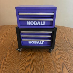 May include: Two-piece purple and black tool chest with the brand name "KOBALT" in white lettering. The top chest has two drawers, and the bottom chest has three drawers and wheels for mobility. The tool chests are sitting on a wooden surface.