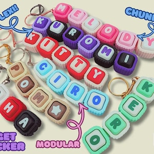May include: A collection of modular fidget clicker keychains made of chunky, colorful square blocks with letters spelling out names like Melody, Kuromi, Kitty, Ciro, and Kerokerokeroppi. Each block features a tactile, pressable surface.