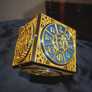 May include: A cubic object featuring intricate gold-colored filigree panels over a deep blue circular base. The blue face displays a central geometric star surrounded by eight distinct gold symbols arranged in a circular pattern on a flat surface.
