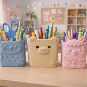 Peut inclure: Trois organisateurs de bureau en forme d'animaux, de couleurs bleu clair, beige et rose. Le support bleu représente un hibou, le beige un ours avec une empreinte de patte, et le rose un lama avec un collier. Ils contiennent divers stylos et ciseaux.