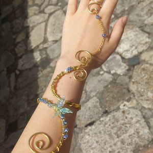Pode incluir: Uma braçadeira de arame dourado com padrões de metal em espiral, pequenas contas azuis e um pingente central de borboleta iridescente. Uma delicada corrente dourada conecta a braçadeira a um anel de arame dourado no dedo médio.