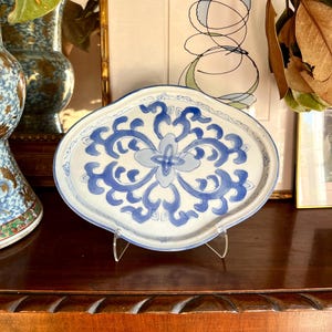 May include: An oval-shaped, white ceramic serving dish with a blue floral design. The dish is supported by a clear acrylic stand and sits on a dark wooden surface. The floral pattern is centered and symmetrical.