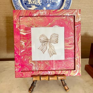 May include: A square pink and gold marbled picture frame with a watercolor bow illustration. The frame is displayed on a small bamboo easel. Decorative plates with blue and white patterns are in the background.