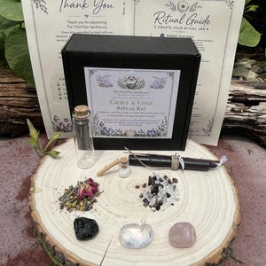 Może przedstawiać: Czarne pudełko z etykietą Grief & Loss Ritual Kit, dwie drukowane instrukcje, szklana fiolka, mała buteleczka z korkiem, czarna świeca, suszone zioła, małe ciemne i jasne kamienie oraz trzy większe polerowane kamienie na drewnianym plastrze.