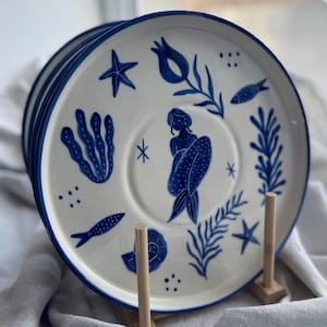 May include: A stack of white ceramic plates with a blue rim and hand-painted nautical designs. Each plate features a mermaid, starfish, fish, and seaweed in blue. The plates are displayed on wooden stands.