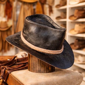 May include: A black leather hat with a light brown braided band, displayed on a wooden hat stand. The hat has a wide brim and a slightly curved crown. Other hats are visible in the background, suggesting a hat shop.