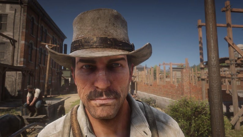 May include: A close-up shot of a man wearing a weathered, light brown hat with a dark band. He has a mustache and is looking directly at the camera. The background shows a building and a street, suggesting a Western theme.
