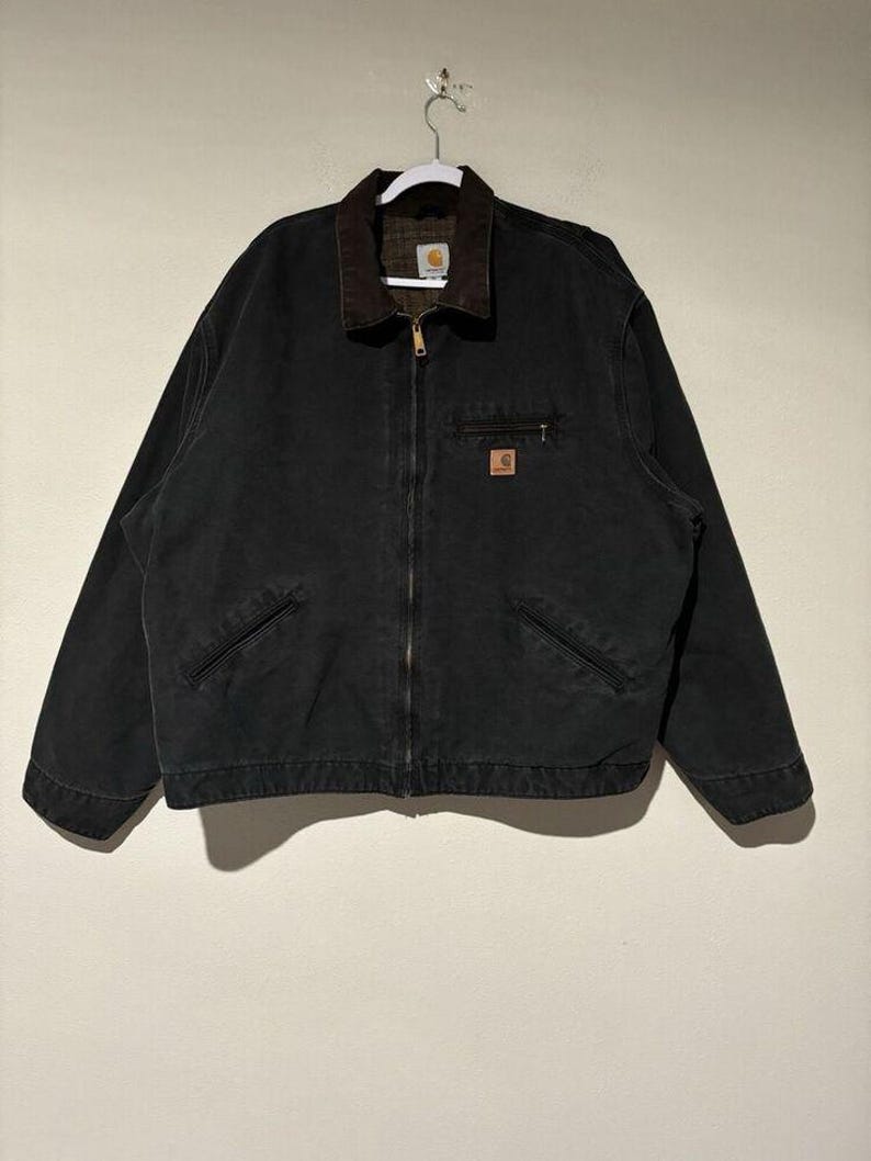 May include: Black work jacket with a brown corduroy collar and a full-length zipper. The jacket has two front pockets and a small square logo patch on the chest. The jacket is hanging on a white hanger.