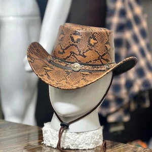 Pode incluir: Um chapéu de cowboy com padrão de pele de cobra castanho e preto, apresentando uma faixa entrançada castanha escura com um emblema metálico circular prateado. O chapéu inclui um cordão fino para o queixo com um fecho ajustável de plástico.