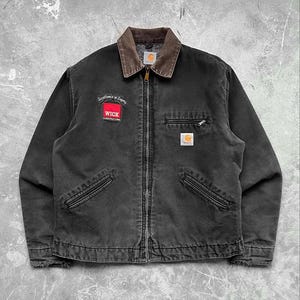 May include: Black work jacket with a brown corduroy collar and a full-length zipper. Features a red and white patch that reads "WICK CONSTRUCTION" and a small Carhartt logo patch. Includes two front pockets and a chest pocket.
