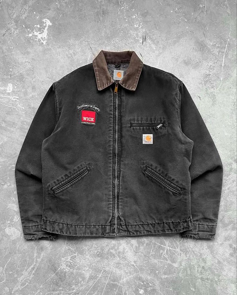 May include: Black work jacket with a brown corduroy collar and a full-length zipper. Features a red and white patch that reads "WICK CONSTRUCTION" and a small Carhartt logo patch. Includes two front pockets and a chest pocket.