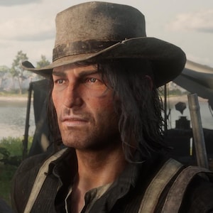 May include: A man wearing a weathered, gray cowboy hat with a dark band. He has long, dark hair and is dressed in a dark shirt with suspenders. The image is a medium shot, with a natural outdoor background.
