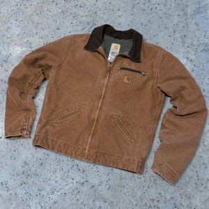 May include: A brown Carhartt jacket with a dark brown corduroy collar and a full zipper. The jacket has two front pockets, a small chest pocket, and the Carhartt logo patch. The jacket is made of a durable, workwear-style material.