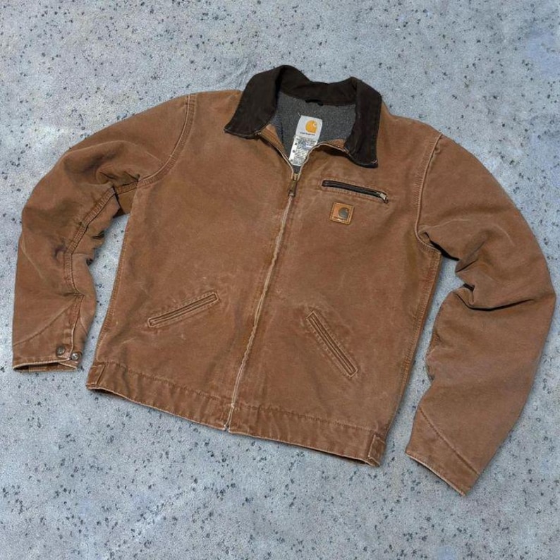 May include: A brown Carhartt jacket with a dark brown corduroy collar and a full zipper. The jacket has two front pockets, a small chest pocket, and the Carhartt logo patch. The jacket is made of a durable, workwear-style material.