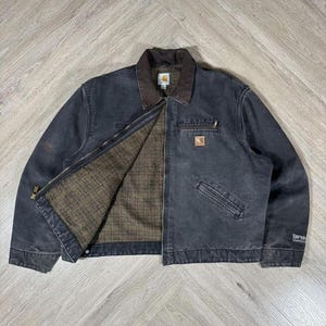May include: A dark gray Carhartt jacket with a brown collar and interior lining. The jacket has a zipper closure, multiple pockets, and a small logo patch on the chest. The jacket is made of durable material and is suitable for work or casual wear.
