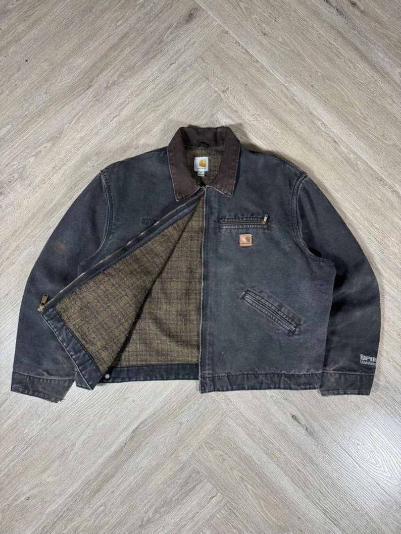 May include: A dark gray Carhartt jacket with a brown collar and interior lining. The jacket has a zipper closure, multiple pockets, and a small logo patch on the chest. The jacket is made of durable material and is suitable for work or casual wear.