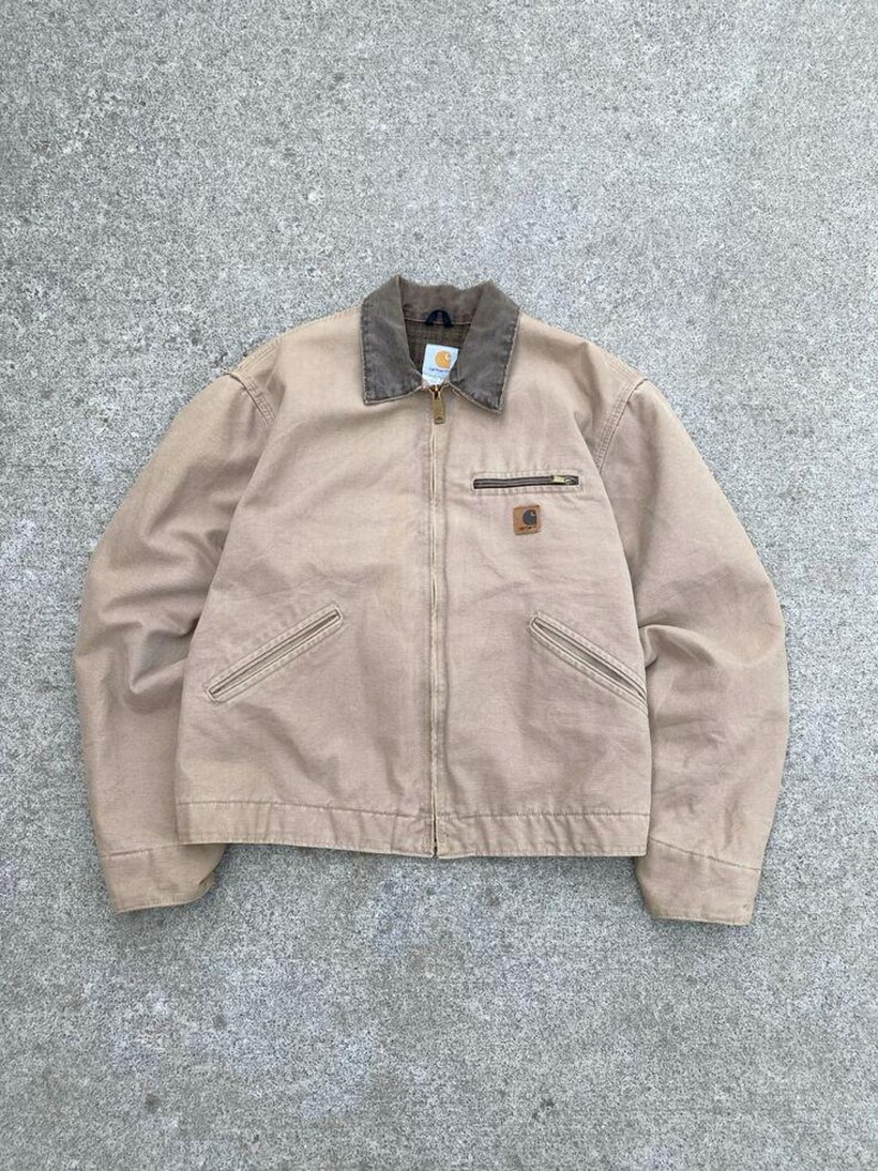 May include: A tan work jacket with a brown collar and a zipper closure. The jacket has two front pockets and a small logo patch on the chest. The jacket is made of a durable, heavy-duty fabric.