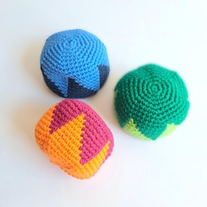 Peut inclure: Trois balles de jonglage rondes au crochet avec des motifs géométriques. Une balle est bleue et bleu foncé, une est verte et vert clair, et une est rose et orange. Chaque balle présente une surface texturée et cousue en fil.