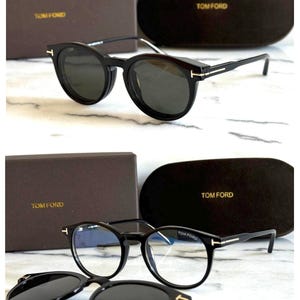 Puede incluir: Gafas de sol Tom Ford negras con estuche y caja a juego. Las gafas tienen una montura redonda con lentes oscuras y un acento dorado en las patillas. El estuche y la caja son marrones con el logo de Tom Ford en dorado.