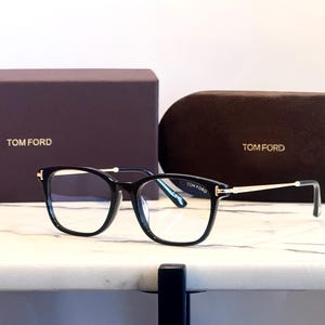 May include: Black framed eyeglasses with gold-toned accents and the brand name "TOM FORD" on the temple. The glasses are displayed with a matching case and box, both bearing the brand name.
