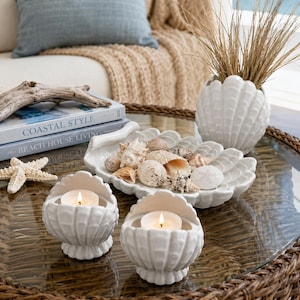 May include: A coastal-themed arrangement featuring white seashell-shaped candle holders with lit candles, a seashell-shaped dish filled with seashells, and a seashell vase with dried grass. The scene is set on a glass-topped table.