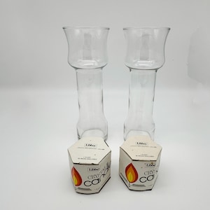 May include: Two clear glass Libbey candle holders with flared tops and cylindrical bases. Each holder is accompanied by a hexagonal cardboard box labeled Libbey Crystal Candle, featuring a flame graphic and text indicating one float and 25 wicks.