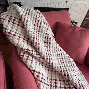 May include: A handmade crochet blanket featuring a classic granny square pattern. The textile is primarily off-white with alternating rows of burnt orange and dark purple yarn accents, creating a textured, repeating geometric design throughout.