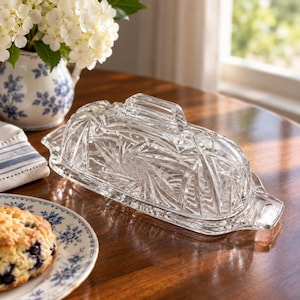 May include: A clear glass butter dish with a rectangular base and a domed lid featuring a faceted starburst pattern and a central handle. The dish has integrated side handles and a smooth, reflective surface that catches the light.