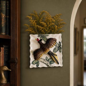 May include: A square ceramic wall pocket featuring a relief of a pheasant in flight with brown, black, and yellow feathers. The white background is decorated with green leaves and branches. A cluster of dried yellow flowers sits inside the pocket.