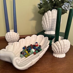 May include: A collection of white ceramic seashell-shaped decor. Includes a large serving dish filled with marbles, two candle holders with blue candles, and a vase with greenery and two candle holders with green candles.