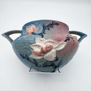 May include: A ceramic bowl with two handles featuring a relief design of white magnolia flowers and dark brown branches. The surface has a blended color gradient transitioning from blue on the left to a muted reddish-pink on the right side.