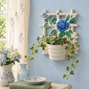May include: A cream-colored ceramic wall planter featuring a lattice grid design with a sculpted blue rose and green leaves. A white pot is attached to the center, containing a trailing green ivy plant with variegated leaves and light edges.