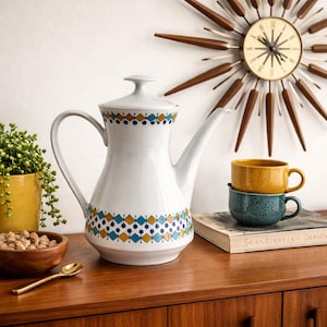 May include: White ceramic coffee pot with a geometric pattern in blue, yellow, and brown. A gold spoon, a wooden bowl, and two stacked mugs sit on a wooden surface. A mid-century modern clock is in the background.