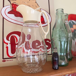 May include: A white ceramic mug with a black handle and the text "But First, Coffee" in a handwritten style. The mug is on a white surface. The mug is approximately 4 inches tall.