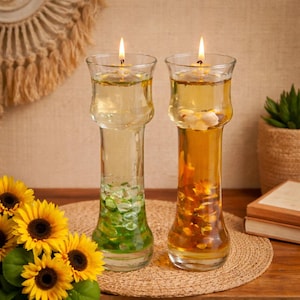 May include: Two glass oil candles with lit wicks. One candle has green stones, the other has orange stones. The candles are on a woven mat with sunflowers in the foreground. The background includes a woven wall hanging and a potted plant.