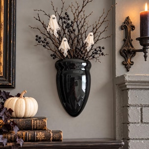 May include: A glossy black wall-mounted vase containing dark brown twigs and black berries, accented with three small white ghost figures draped in fabric. The vase has a smooth, tapered shape and is mounted on a flat, neutral-colored wall.