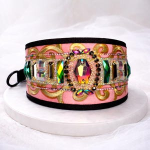 May include: Pink dog collar with gold filigree and a black trim. The collar is embellished with a row of rectangular and faceted gemstones in shades of green, clear, and iridescent. A large, multi-faceted gem is centered on the collar.